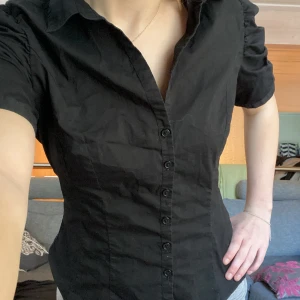 Svart kortärmad skjorta från H&M - Säljer en svart skjorta från H&M med korta puffärmar och klassisk krage. Skjortan har knappar framtill och figurnära passform som ger en snygg siluett. Perfekt till många olika outfits!
