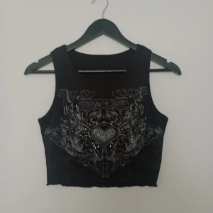 Svart croppad linnetopp med tryck - Säljer en svart croppad linnetopp från Romwe med coolt tryck och glittriga detaljer framtill. Toppen är ärmlös och har ett edgy motiv med hjärta och kedjor. Perfekt för att lyfta din outfit!