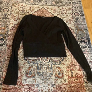 Svart omlottopp med lång ärm - Säljer en svart omlottopp med lång ärm och croppad passform. Toppen har en djup v-ringning och är perfekt att styla med höga jeans eller kjol. Enkel och stilren design som passar till många tillfällen.
