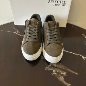 Grå sneakers i mocka från Selected Homme - Säljer ett par stilrena grå sneakers från Selected Homme i mjuk mocka. Skorna har gröna snörning, vit platt sula och en klassisk rund tå. Perfekta för dig som gillar en enkel och clean look.