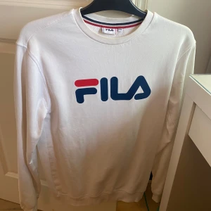 Vit sweatshirt från FILA - Vit sweatshirt från FILA med stor logga framtill i blått och rött. Perfekt för en sportig och avslappnad stil. Sparsamt använd😁storlek xs. Perfekt  till ett småsyskon kanske☺️