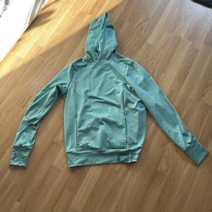 Ljusgrön/turkos hoodie från Craft - Säljer en ljusgrön/turkos hoodie från Craft med långa ärmar och huva. Tröjan har tumhål vid ärmsluten och diskret logga på nederkanten. Perfekt för dig som gillar sportig stil och vill ha något bekvämt och snyggt.