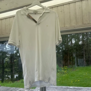Vit piké t-shirt från Burberry - Vit piké t-shirt från Burberry med klassisk krage och korta ärmar. Typisk Burberry-rutig detalj på insidan av kragen. Perfekt för en stilren och avslappnad look.
