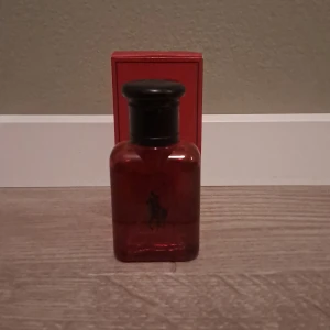 Röd parfymflaska från Ralph Lauren Polo - En stilren parfymflaska i rött glas med svart kork från Ralph Lauren Polo. Flaskan har den klassiska Polo-loggan framtill och kommer i en matchande röd förpackning. Perfekt för dig som gillar eleganta och tidlösa dofter.