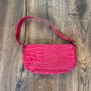 Röd axelväska i krokodilpräglad syntet - Cool röd axelväska med krokodilpräglat mönster och enkel axelrem. Perfekt för att lyfta vilken outfit som helst med en färgklick och trendig look. Väskan har en stilren form och är lagom stor för det viktigaste.