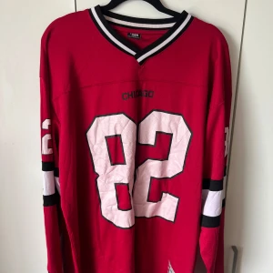Röd Chicago-tröja med nummer 82 - Säljer en röd långärmad Chicago-tröja med nummer 82 i vitt och svart på både fram- och baksidan. Tröjan har svarta och vita detaljer på ärmarna och en vit-svart krage. Perfekt för dig som gillar sportig stil.