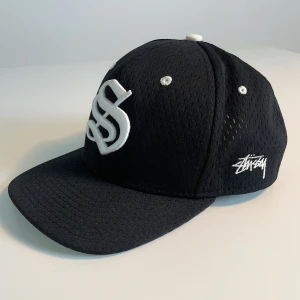 Stüssy keps - Svart keps från Stüssy med vit broderad logga framtill och på sidan. Kepsen är tillverkad i mesh-liknande polyester och har justerbar snapback.