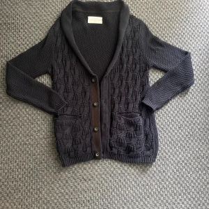 Mörkblå stickad kofta från These Glory Days - Säljer en mörkblå stickad kofta från These Glory Days i storlek medium. Koftan har ett snyggt strukturmönster, två framfickor och knappar framtill. Perfekt för lager-på-lager och har en klassisk krage.