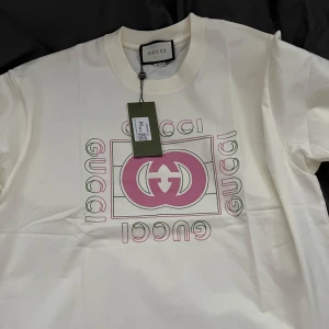 Gucci T-Shirt - Säljer en vit t-shirt från Gucci med ikonisk rosa logga och text framtill, den sitter lite stor i storleken men det är så den ska göra så den passar M/L. T-shirten har klassisk passform och rund halsringning. För fler bilder/disskusion skriv pm, inga byten och oseriösa köpare tack!