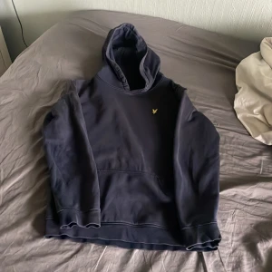 Mörkblå hoodie från Lyle & Scott Junior - Säljer en mörkblå hoodie från Lyle & Scott Junior med klassisk huva och magficka. Tröjan har en liten gul logga på bröstet och är perfekt för en avslappnad stil. Mjuk och bekväm, passar till det mesta.