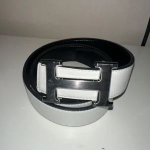 Vitt skärp med H-spänne - Snyggt vitt bälte från Her-mès med ett stort, silverfärgat H-spänne. Bältet är gjort i skinn och har en stilren design med svart insida. Perfekt accessoar för att lyfta din outfit. 100cm