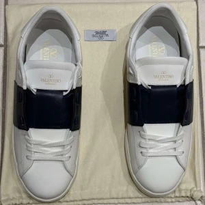 Valentino Garavani vita sneakers med svart band - Säljer ett par snygga vita sneakers från Valentino Garavani med ett brett svart band över mitten. Skorna har rund tå, platt sula och är tillverkade i skinn. Klassisk och stilren design med diskret logga på plösen och insidan.