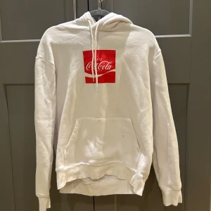 Vit hoodie från H&M x Coca-Cola - Vit hoodie från H&M i samarbete med Coca-Cola. Stor röd Coca-Cola-logga framtill och flera olika Coca-Cola-loggor på ryggen i olika språk. I strl M