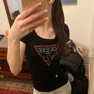 Svart t-shirt från Guess med röd logga - Svart t-shirt från Guess med ikonisk röd triangel-logga framtill. T-shirten har rund halsringning och korta ärmar. Perfekt för en trendig och avslappnad look. Storleken är ungefär xs-s🥰🥰smutsen är i speglet