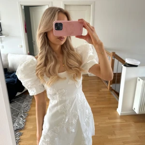 Vit klänning med puffärmar och blommiga detaljer - Säljer en vit klänning med stora puffärmar och vackra blommiga broderier. Klänningen har en figurnära passform och är perfekt för speciella tillfällen. Materialet känns lyxigt och ger en elegant look.