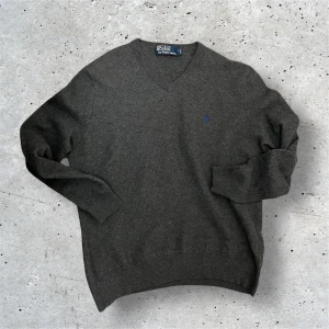 Polo Ralph Lauren Stickad Tröja - Polo Ralph Lauren ulltröja – 99kr Klassisk mörkgrå stickad tröja i 100% ull från Polo by Ralph Lauren. V-ringad modell med diskret blå logga på bröstet.  Tröjan är i fint skick i övrigt, men priset är nedsatt till 69kr pga ett litet hål vid halsringningen (se bild). Står att det är storlek L men passar S då den krympt i tvätten. Perfekt för dig som gillar second hand-fynd och kan sy eller bära den som den är!  Storlek: S (men passar även XS beroende på passform) Material: 100% ull