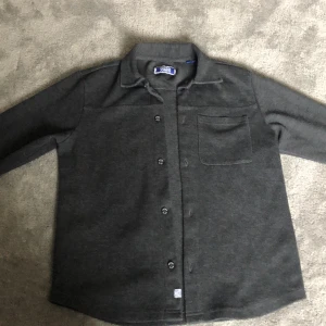 Mörkgrå skjorta från Jack & Jones - Säljer en mörkgrå skjorta från Jack & Jones med klassisk krage, bröstficka och knäppning framtill. Skjortan har långa ärmar och är tillverkad i mjukt material som passar perfekt till en avslappnad stil.