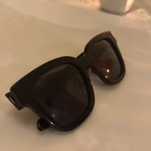 Gucci solglasögon  - Stilrena svarta solglasögon från Gucci med markerad båge och klassisk GG-logga i guld på sidorna. Glasögonen har en fyrkantig form och mörka linser, perfekt för en trendig look. Ordinarie pris 4600. Kvitto finns tyvärr inte, trodde jag inte skulle sälja