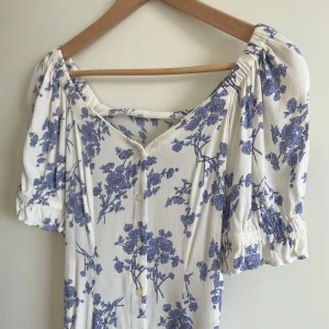 Vit och blå blommig klänning från Mango - Superfin vit klänning från MNG med blått blommönster. Klänningen har korta puffärmar, knappar framtill och en lätt v-ringad halsringning. Perfekt för sommardagar och har en avslappnad passform.