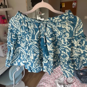 Kort blå kjol med tofsar - Jag säljer nu denna superfina kjolen med volanger och vita mönster, detaljerade tofsar(en av pärlslingorna runt har åkt av) annars inga defekter!! Från Gina Young och sälj inte längre!!💙💙