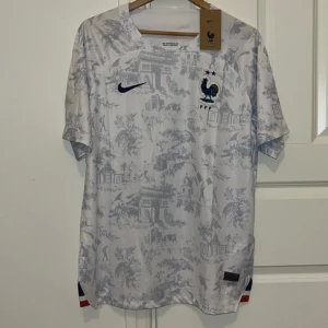 Frankrike fotbollströja Nike - Säljer en officiell Frankrike fotbollströja från Nike i vitt med subtilt grått mönster av franska landmärken. Tröjan har korta ärmar, broderad tupp-logga och Nike swoosh på bröstet. Tillverkad i Dri-FIT material för bästa komfort.