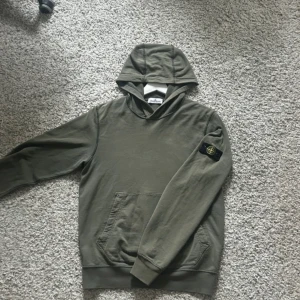 Stone Island hoodie - Säljer en olivgrön hoodie från Stone Island Junior med klassisk patch på vänster ärm. Tröjan har huva och magficka, samt ribbade muddar vid ärmslut och nederkant. Perfekt för en avslappnad och stilren look. Skriv för minsta frågor eller funderingar 