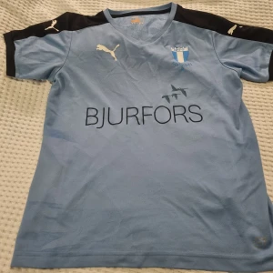 MFF fotbollströja från Puma - Säljer en ljusblå Malmö FF fotbollströja från Puma med korta ärmar och svarta axelpartier. Tröjan har klubbens emblem på bröstet och sponsorloggan 'Bjurfors' tryckt på framsidan. Perfekt för alla MFF-fans! 💙