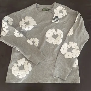 Grå långärmad tröja med vita blommor från Denim Tears - Säljer en grå långärmad tröja från Denim Tears med vita blommor tryckta över hela tröjan. Tröjan har rund halsringning och är perfekt för dig som gillar unika prints. Märkt med Dover Street Market-tag. Den är oanvänd då jag inte tyckte om den och är i storlek M. Pris går även att diskuteras!