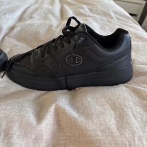 Svarta sneakers från Champion - Svarta sneakers från Champion med klassisk design och snörning. Skorna har rund tå, helsvart ovandel och sula samt diskret Champion-logga på sidan. Perfekta för en stilren vardagslook.