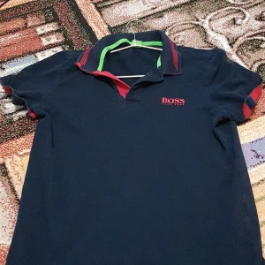 Mörkblå pikétröja från Hugo Boss - Snygg mörkblå pikétröja från Hugo Boss med röd logga på bröstet. Tröjan har en klassisk krage med gröna och röda detaljer. Perfekt för en stilren look.