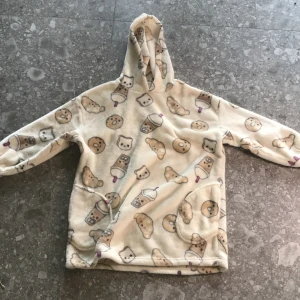 Beige fleecehoodie med söta mönster från Kappahl - Supermjuk och mysig beige hoodie i fleece från Kappahl. Tröjan har huva och är täckt av gulliga mönster med katter, glassar, donuts och croissanter. Perfekt för att hålla sig varm och bekväm hemma eller ute.