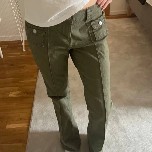 Olivgröna cargopants med fickor - Snygga olivgröna cargopants med raka ben och stora fickor framtill med knappar. Byxorna har hög midja och markerade sömmar längs benen för en cool look. Perfekta för dig som gillar utility-stil.