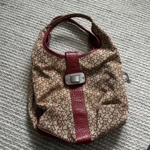 Beige och röd axelväska med monogram - Snygg axelväska från DKNY med monogrammönster i beige och rött. Väskan har röda skinnremmar och glera fack! Perfekt för att bära dina dagliga nödvändigheter med stil.