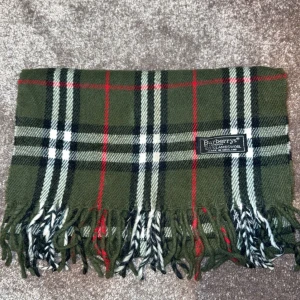  Burberry halsduk - Snygg grön rutig halsduk från Burberry med klassiskt mönster i svart, vitt och rött. Halsduken har fransar i ändarna och är perfekt för att hålla dig varm och stilren under kyliga dagar.