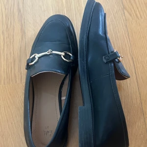 Svarta loafers med guldfärgad kedjedetalj - Stilrena svarta loafers från H&M med guldfärgad kedjedetalj framtill. 