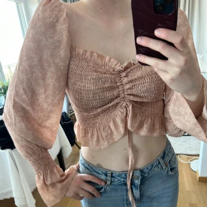 Beige croppad blus med volang och knyt - Säljer en söt beige/ljusrosa croppad blus med små vita blommor, volangkant och knyt framtill. Blusen har långa puffärmar med resår vid handlederna och smockad överdel för en tight passform. Perfekt för vår och sommar! Aldrig använd. 