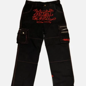 minus two Svarta cargobyxor med röd grafik och fickor - Unika svarta cargobyxor med röda grafiska tryck och illustrationer både fram och bak. Byxorna har stora fickor med dragkedja på sidorna och detaljerade sömmar. Perfekta för dig som vill sticka ut med en edgy streetstyle.