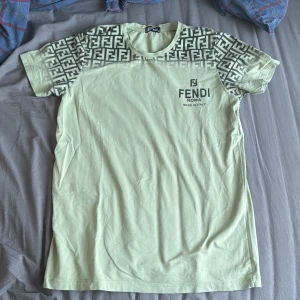  t-shirt från Fendi - Säljer en snygg beige t-shirt från Fendi med korta ärmar och ett coolt mönster på axlarna. T-shirten har Fendi-loggan tryckt på bröstet. Perfekt för dig som vill ha något stilrent men ändå med en touch av lyx.