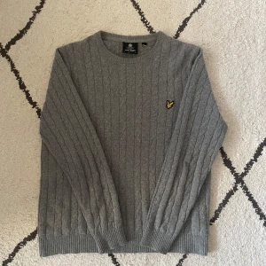 Grå stickad tröja från Lyle & Scott - Säljer en stilren grå stickad tröja från Lyle & Scott med kabelstickat mönster. Tröjan har en rund halsringning och ett litet broderat emblem på bröstet. Perfekt för kyligare dagar.