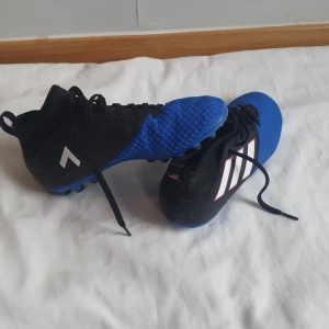 Adidas fotbollsskor blå och svarta - Säljer ett par Adidas fotbollsskor i svart och blått med vita ränder på sidan. Skorna har snörning och mönstrad ovandel för extra grepp. Perfekta för dig som vill ha stilrena och sportiga skor på planen.