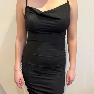Svart långklänning med smala axelband - Elegant svart långklänning med smala axelband och draperad design. Perfekt för en stilren look. Stretchig så passar även större storlekar