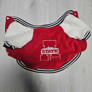 Röd och vit varsityjacka med broderi - Säljer en röd och vit varsityinspirerad jacka till hunden med broderad text 'STATE UNIVERSITY' på ryggen. Jackan har svarta och vita ränder på krage, ärmslut och nederkant samt vita tryckknappar framtill. Mjuk och fluffig känsla, perfekt för en sportig look.