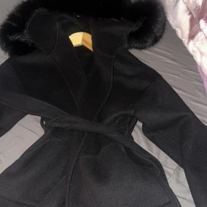 Svart kappa med fuskpäls och bälte - Säljer en svart kappa med stor huva och fuskpälsdetalj runt kanten. Kappan har bälte i midjan för snygg passform och stora fickor framtill. Perfekt för kyliga dagar och ger en stilren look. helt ny användning två gånger. Pris 1800kr kan diskuteras vid snabb köp. Äkta päls. KÖPT DEN FÖR 3800kr