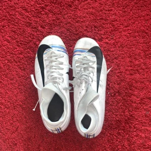 Vita fotbollsskor från Nike med svarta detaljer - Säljer ett par vita fotbollsskor från Nike med svart swoosh och blåa samt gula ränder vid hälen. Skorna har snörning och rund tå. Perfekta för dig som vill sticka ut på planen!
