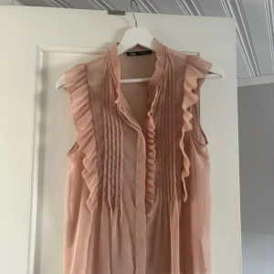 Ljusrosa blus med volang från Zara - Säljer en ljusrosa blus från Zara med volangdetaljer längs ärmhål och framtill. Blusen är ärmlös och har fina veckade detaljer framtill. Perfekt för en somrig och feminin look!
