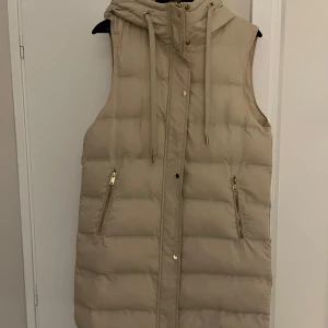 Snygg beige dunväst från Zara - Snygg beige täckväst från Zara med gulddetaljer. Västen har en huva med dragsko och fickor med dragkedja. Jättefin och endast använd ett fåtal gånger då jag tyvärr köpte för stor storlek.