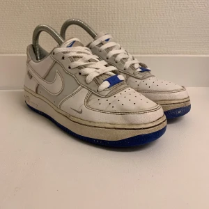 Nike Air Force 1 med blå sula, strlk 36  - Säljer ett par klassiska Nike Air Force 1 sneakers i vitt skinn med grå detaljer och blå sula. Skorna har snörning och rund tå, samt tydlig Nike-logga på sidan och hälen. Perfekta för dig som gillar Stockholm och streetwear stil. (Små skador) på skon, märks knappt.