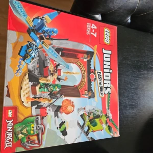 LEGO Juniors Ninjago 10725 - Säljer ett LEGO Juniors Ninjago set, modell 10725. Lådan är röd med färgglada illustrationer av ninjagubbar, svärd, helikopter och tempel. Setet är märkt 'Easy to Build' och passar för barn 4-7 år. Perfekt för unga byggare som gillar action och äventyr.