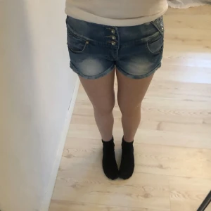 Blå jeansshorts med broderi - Snygga blå jeansshorts med dekorativa knappar framtill. Shortsen har broderade detaljer och nitar på baksidan som ger en unik touch. Perfekta för sommardagar! 💙
