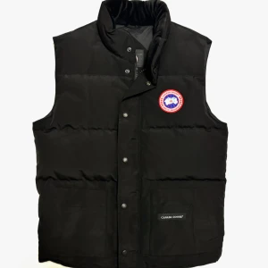 Canada Goose väst  - Säljer en stilren svart dunväst från Canada Goose. Västen har en hög krage och knappar framtill. Perfekt för kyliga dagar när du vill hålla värmen utan att kompromissa med stilen.